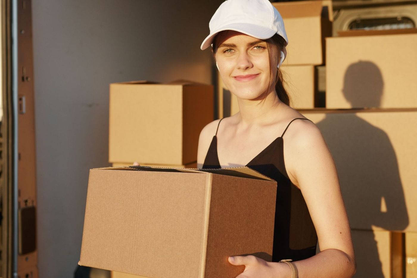 A woman in a white cap holding a cardboard box in front of a van stacked with more boxes