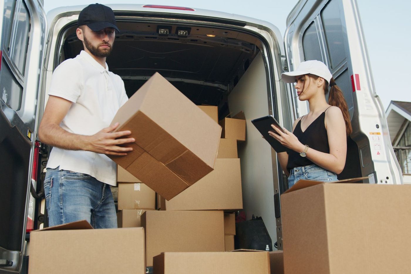 A man lifting a cardboard box at the open rear doors of a van while a woman beside him checks a tablet